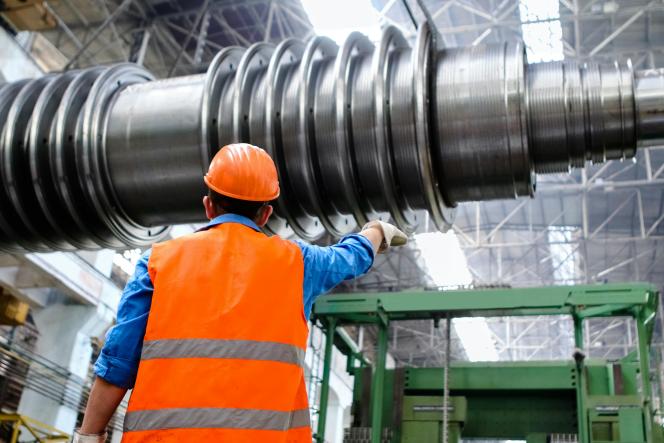 Ein Mann mit Helm und Warnweste deutet auf ein großes Metallteil in einer großen Industrielagerhalle