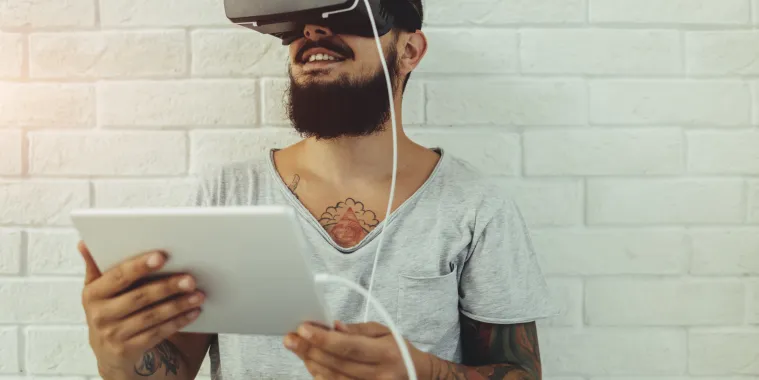 ein junger Mann hält ein Tablet in der Hand und trägt eine VR-Brille.